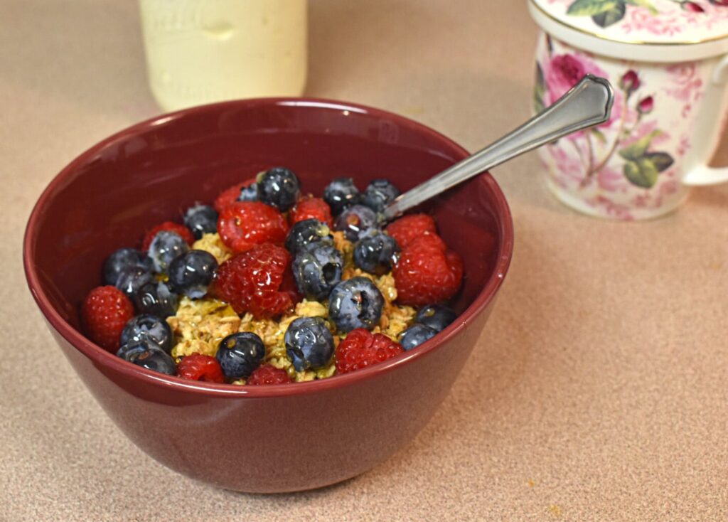 instant pot greek yogurt with blueberries, raspberries, granola, and honey on top in a red bowl with a tea cup covered in roses in the background