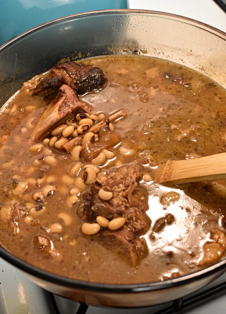 looking down into a clear pot of southern black eyed peas with ham bones and a wooden spoon inside