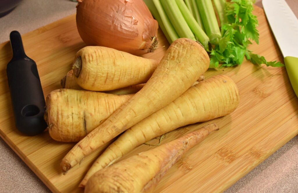 whole parsnips, onion, and celery resting on a wooden cutting board and a knife