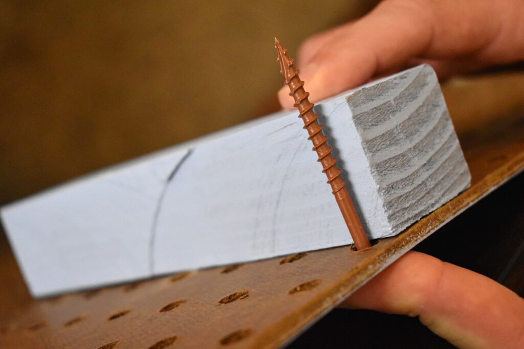 a screw through a peg board and next to a piecer of wood to compare length