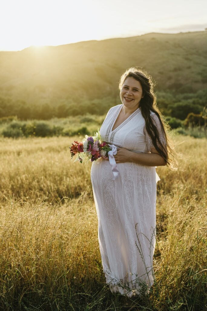 Sophia White a brunette woman in white dress pregnant with twins in a field holding flowers